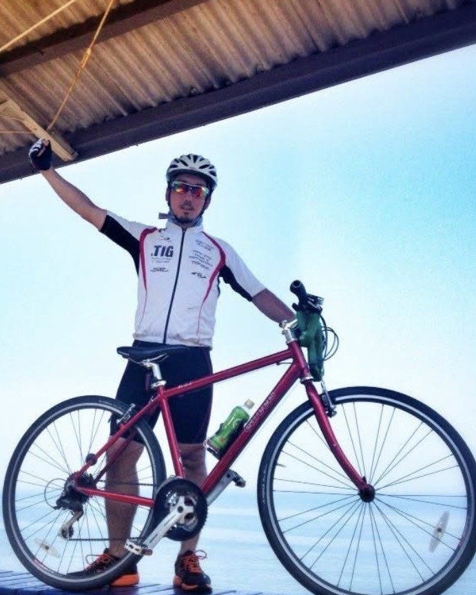 海の見える屋根の下で、自転車の横に立ち片手を上げる石村侑二