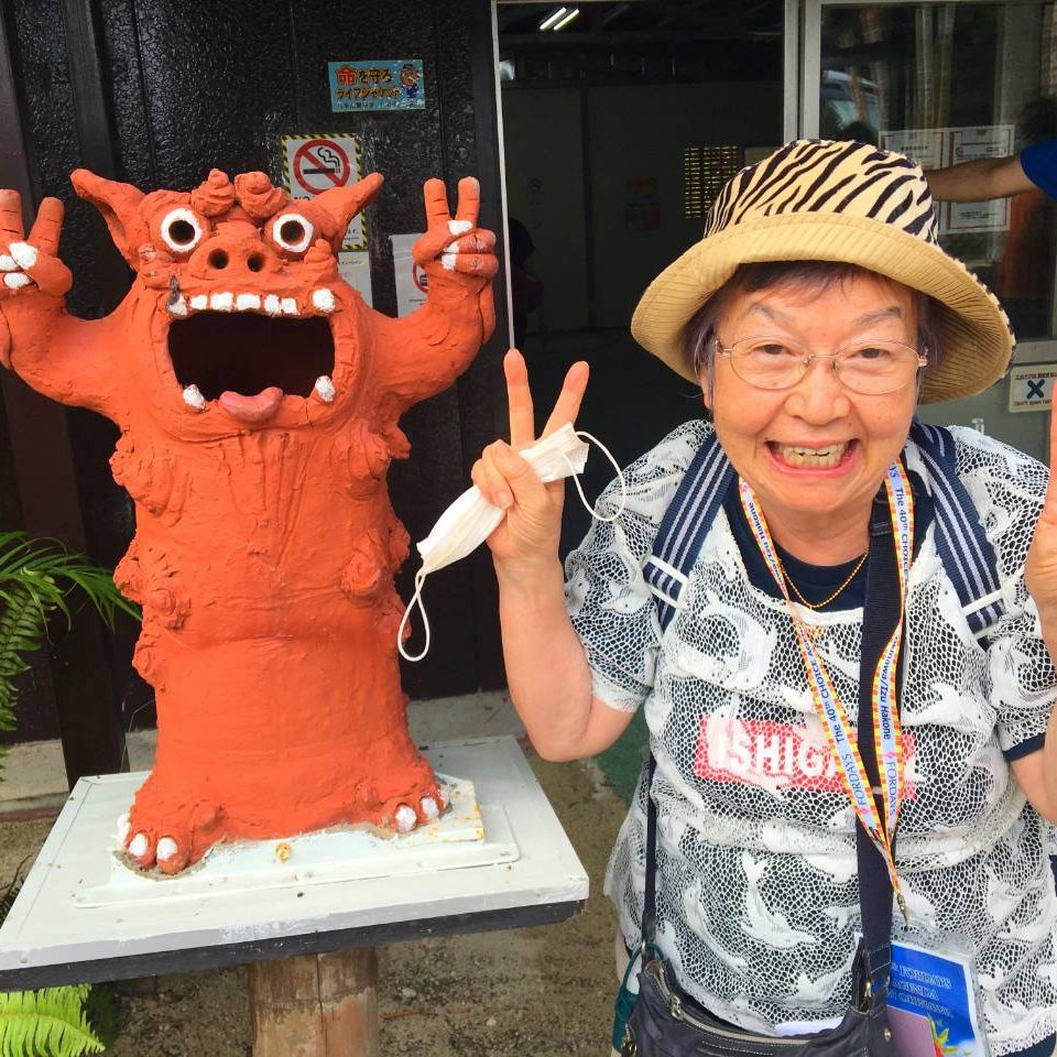 赤いシーサーの像の横で、麦わら帽子をかぶって両手でピースをするますみ