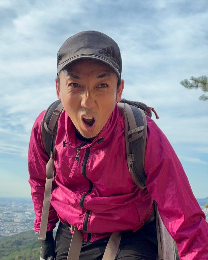 山の上で、ピンクのジャケットにリュックを背負い、口を大きく開けてこちらを見る石村侑二
