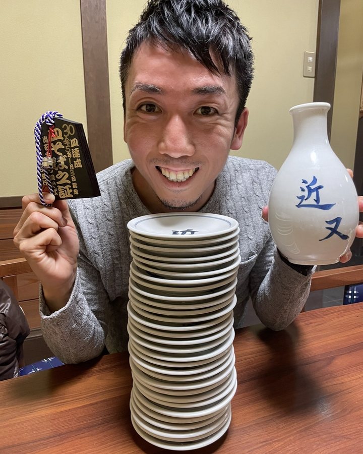 積み上げた皿そばの皿と「30皿達成」の木札を手に笑う石村侑二
